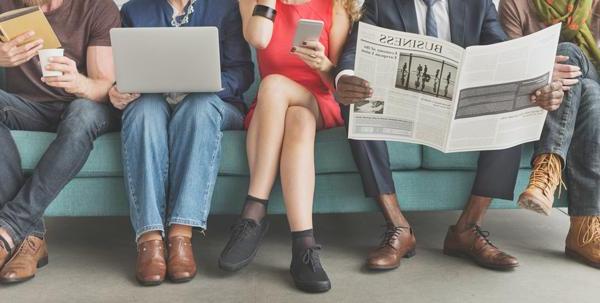 Several people sitting on a couch reading either a newspaper, a book, a smartphone, or a laptop.